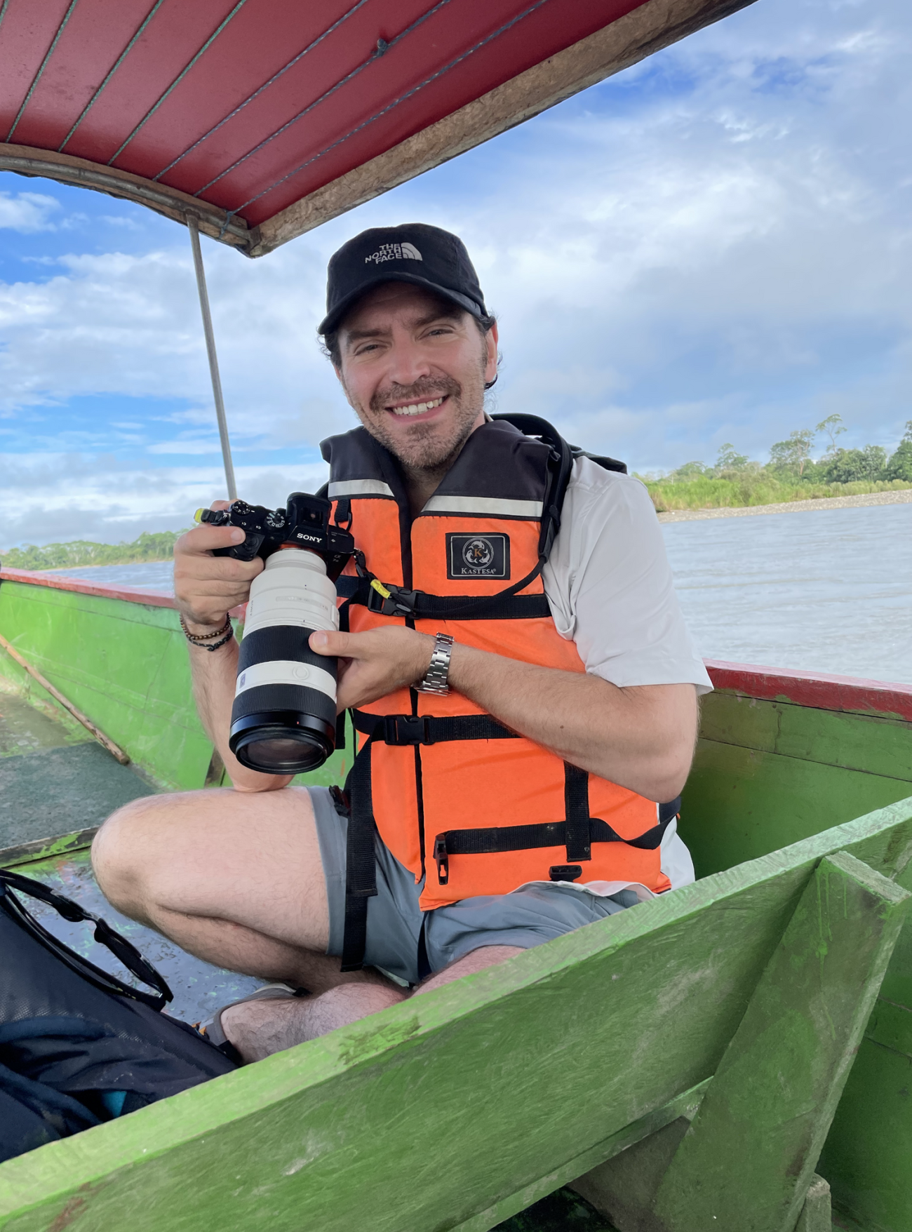 Sitting cross-legged in the bow of a colorful, wooden river boat wearing a orange life jacket and shorts, Sean cradles a Sony with a long telephoto lens.